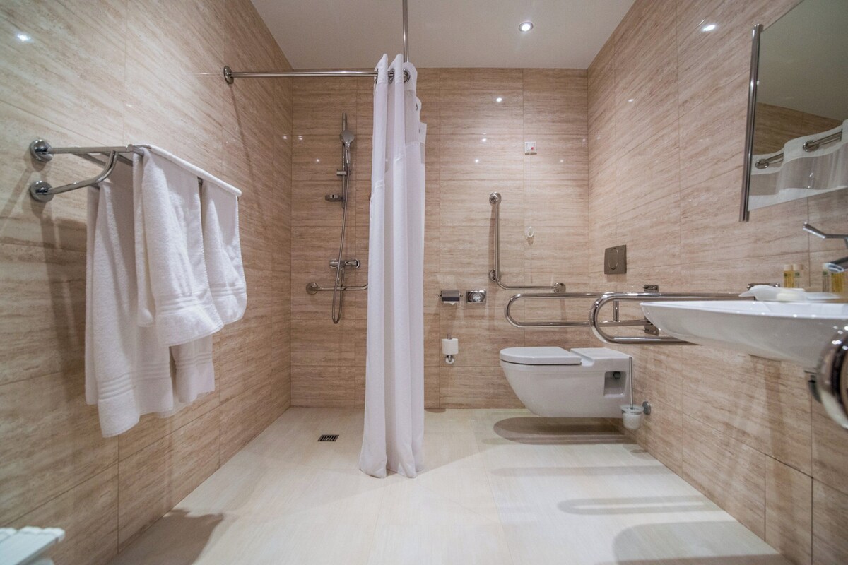 Uma casa de banho moderna com chuveiro, sanita e lavatório, equipada com barras de apoio e secador de mãos.