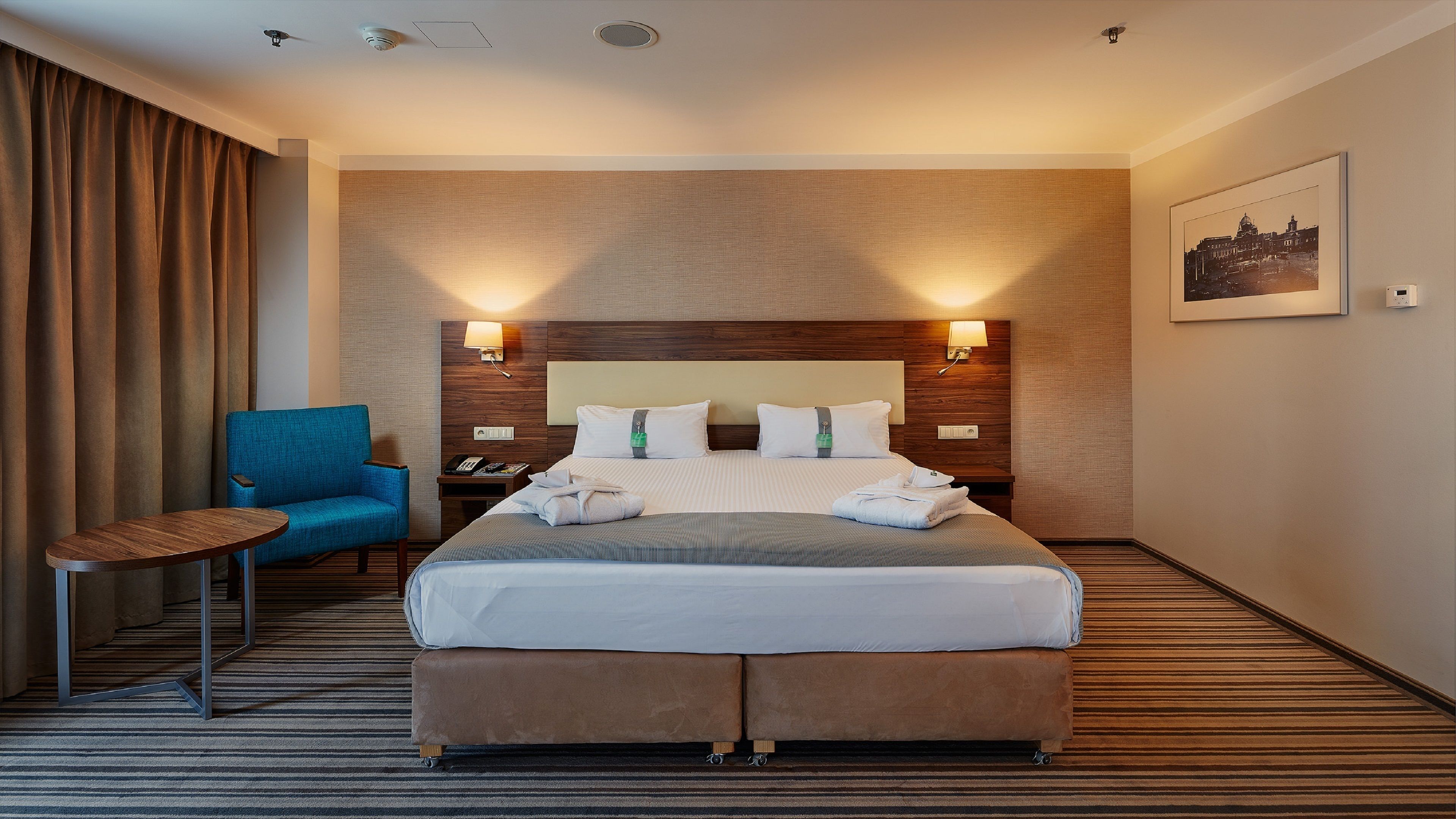 Una habitación de hotel con una cama grande, un sillón azul, una mesita pequeña y una lámpara de pared.