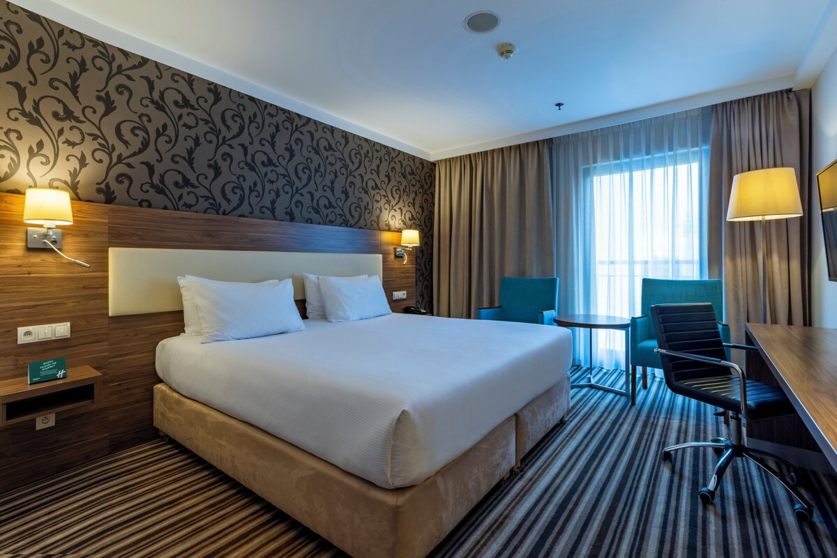 Quarto de hotel com uma cama grande, uma escrivaninha, uma cadeira e uma mesinha.