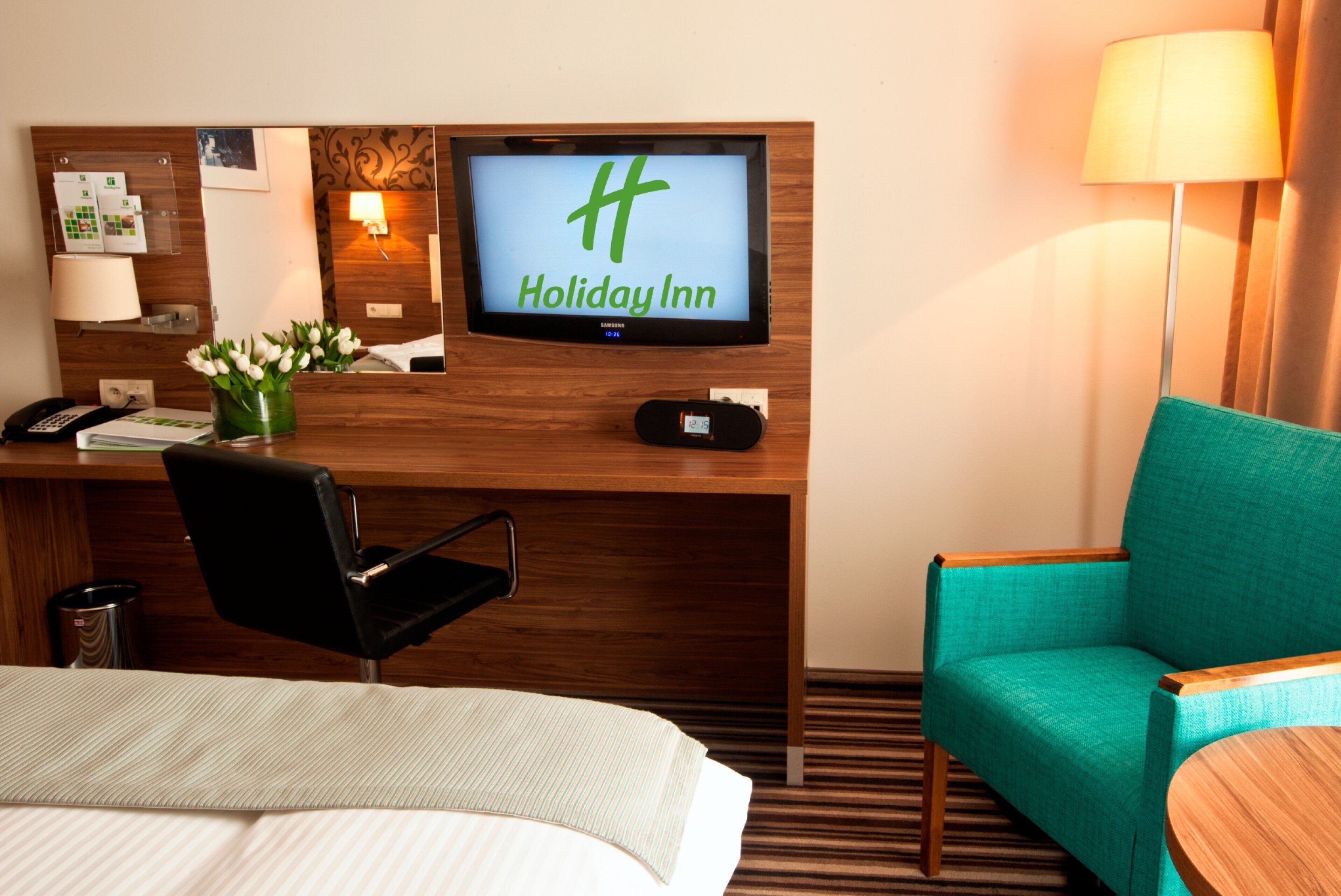 Habitación de hotel con una cama, un escritorio con una computadora, un televisor mostrando 'Holiday Inn', un sillón verde azulado y una lámpara.