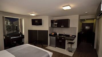A compact hotel room with a bed, a desk, a kitchenette, and a small seating area.