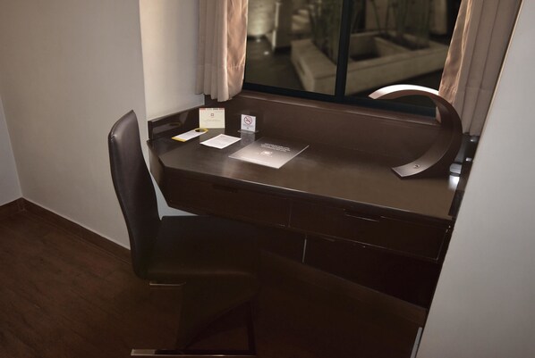 Un escritorio marrón oscuro con una silla, una ventana con cortinas, y una mesita con un cartel y papeles.