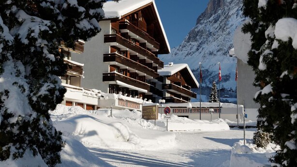 Un hotel con varios balcones, un centro de esquí al fondo y árboles cubiertos de nieve.