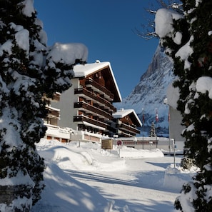 A hotel with multiple balconies, a ski resort in the background, and snow-covered trees. - Residence Hotel & Apartments (Grindelwald)
