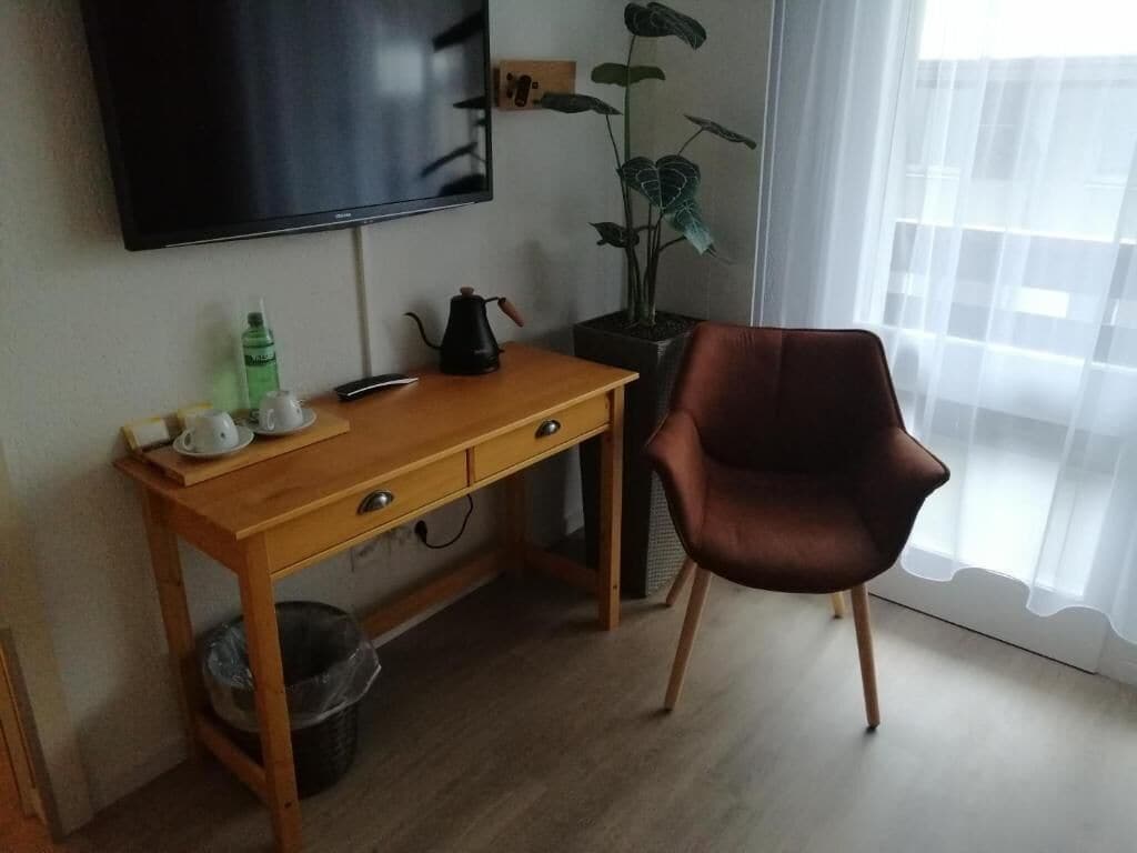 Una habitación con un escritorio de madera, una silla marrón, un televisor montado en la pared, una planta en maceta y una ventana con cortinas traslúcidas.