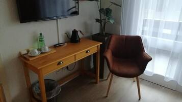 Una habitación con un escritorio de madera, una silla marrón, un televisor montado en la pared, una planta en maceta y una ventana con cortinas traslúcidas.