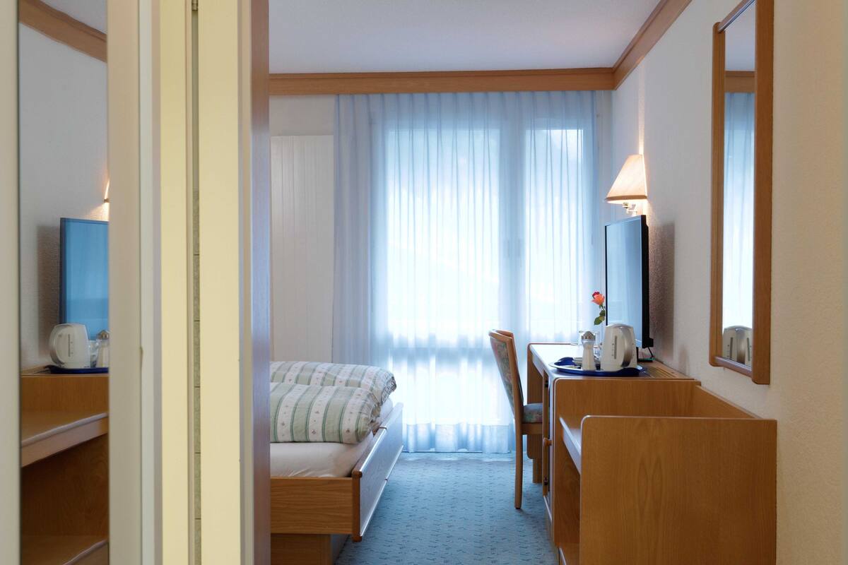 Quarto de hotel com cama, escrivaninha, cadeira, luminária e janela com cortinas leves.