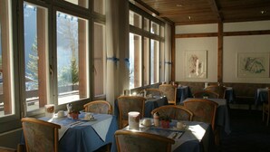 Un restaurante con mesas dispuestas para comer, ventanas amplias con cortinas y obras de arte enmarcadas en las paredes.