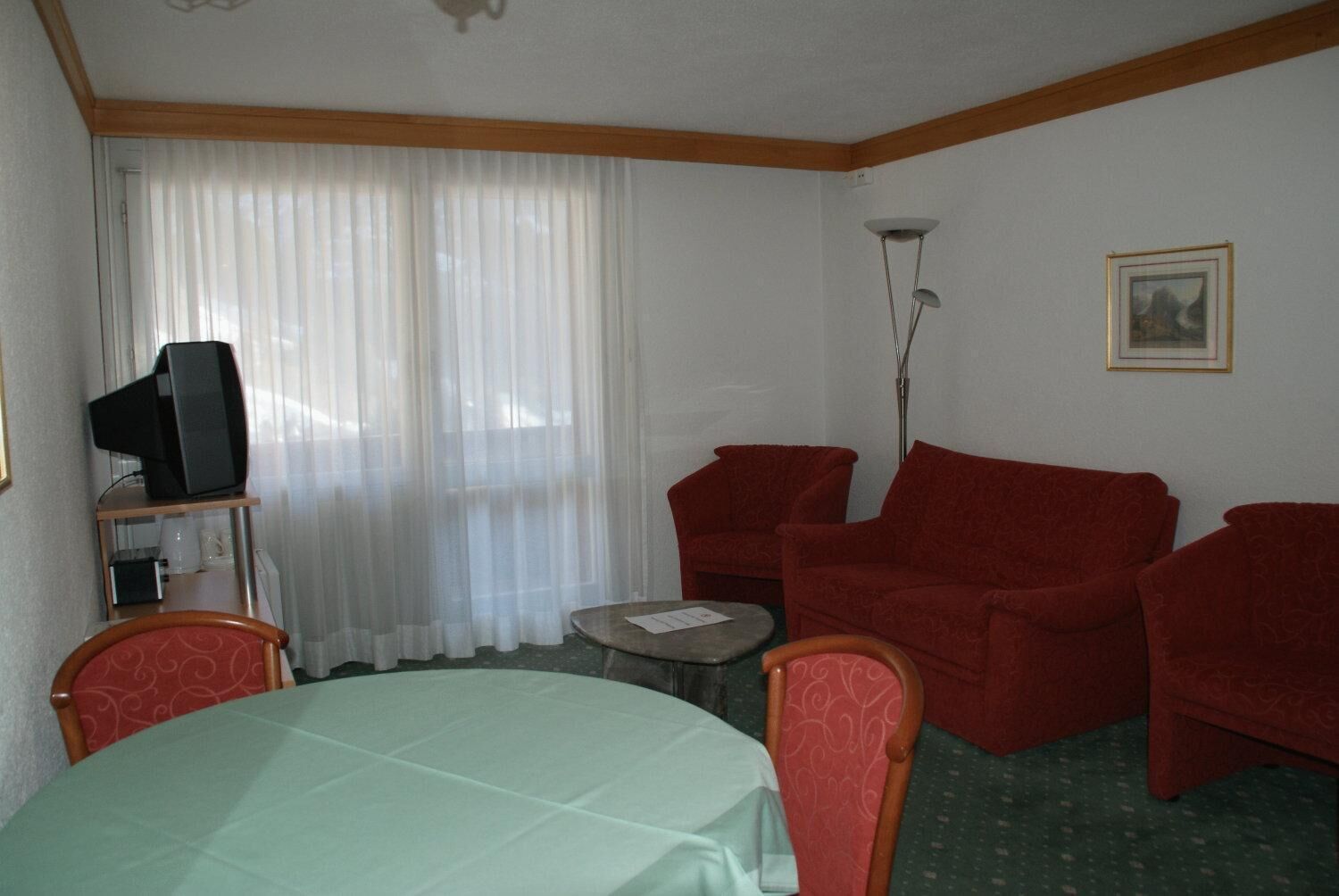 A room with a dining table, two red armchairs, a sofa, a television on a stand, and a floor lamp.