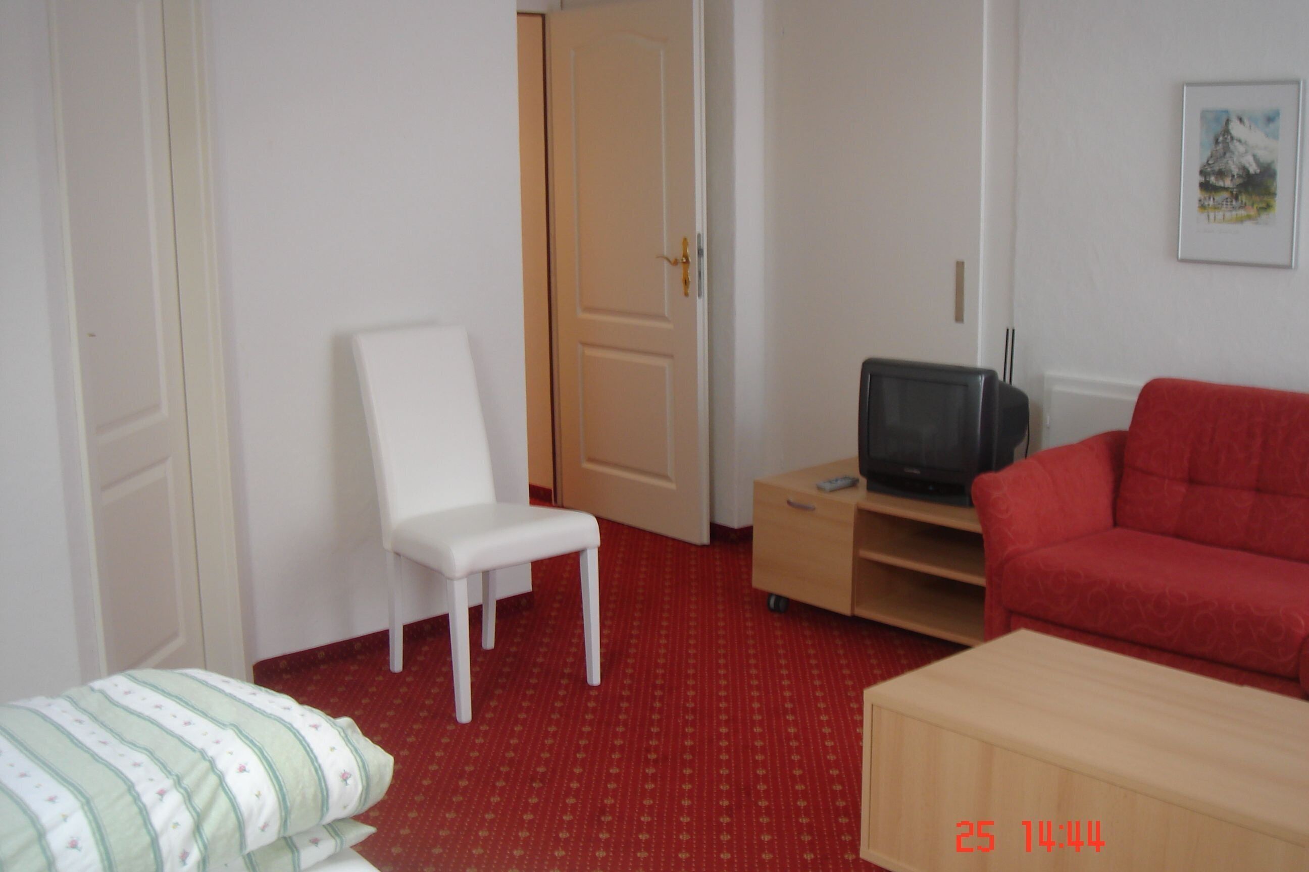 Una habitación con una silla blanca, un sofá rojo, un televisor sobre un mueble de madera y una puerta que da a otra estancia.