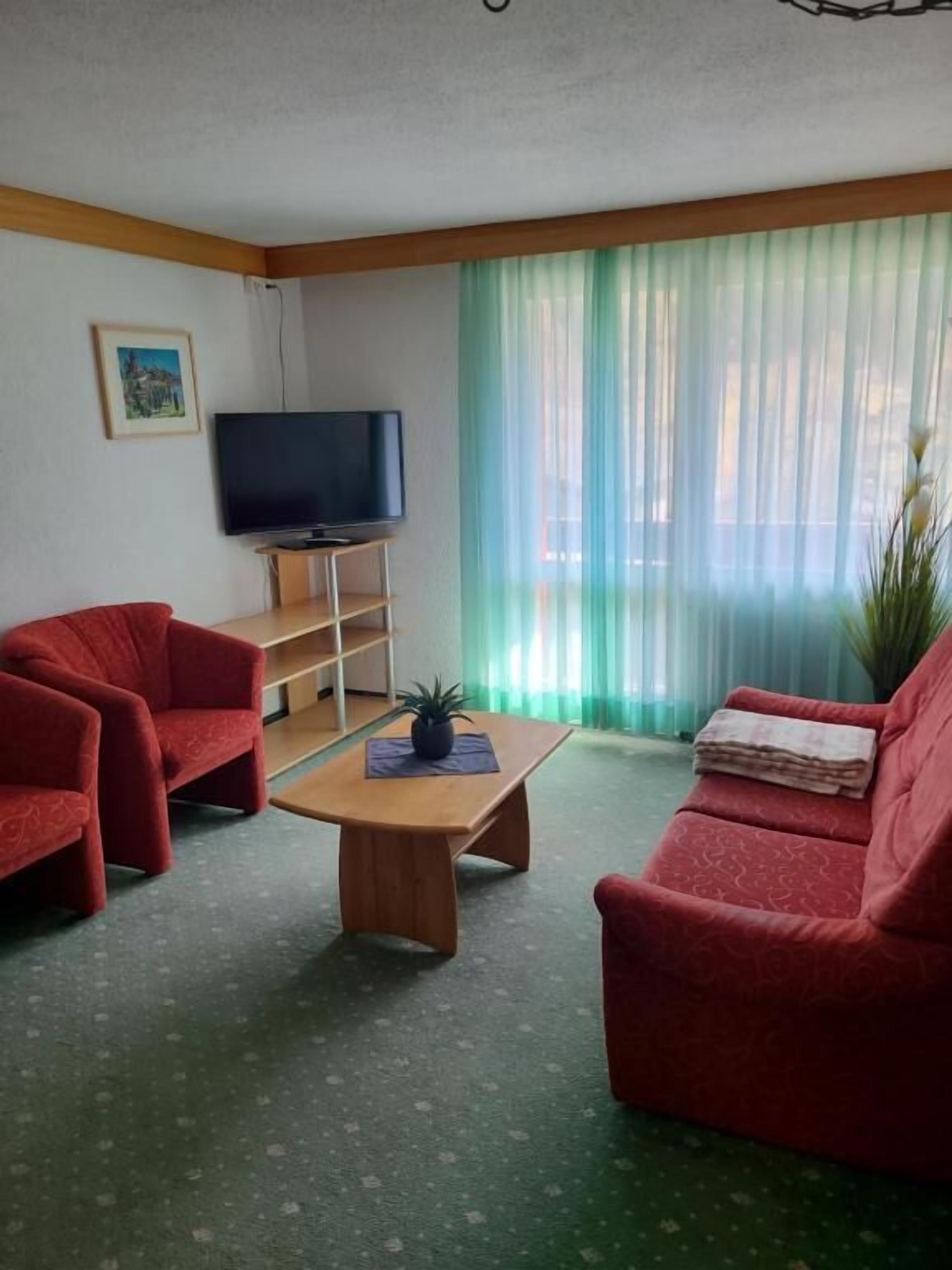 A living room with a television on a stand, two red sofas, a wooden coffee table, and a large window with sheer curtains.