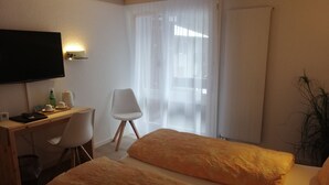 Habitación de hotel con cama, escritorio, silla, televisor y ventana con cortinas traslúcidas.