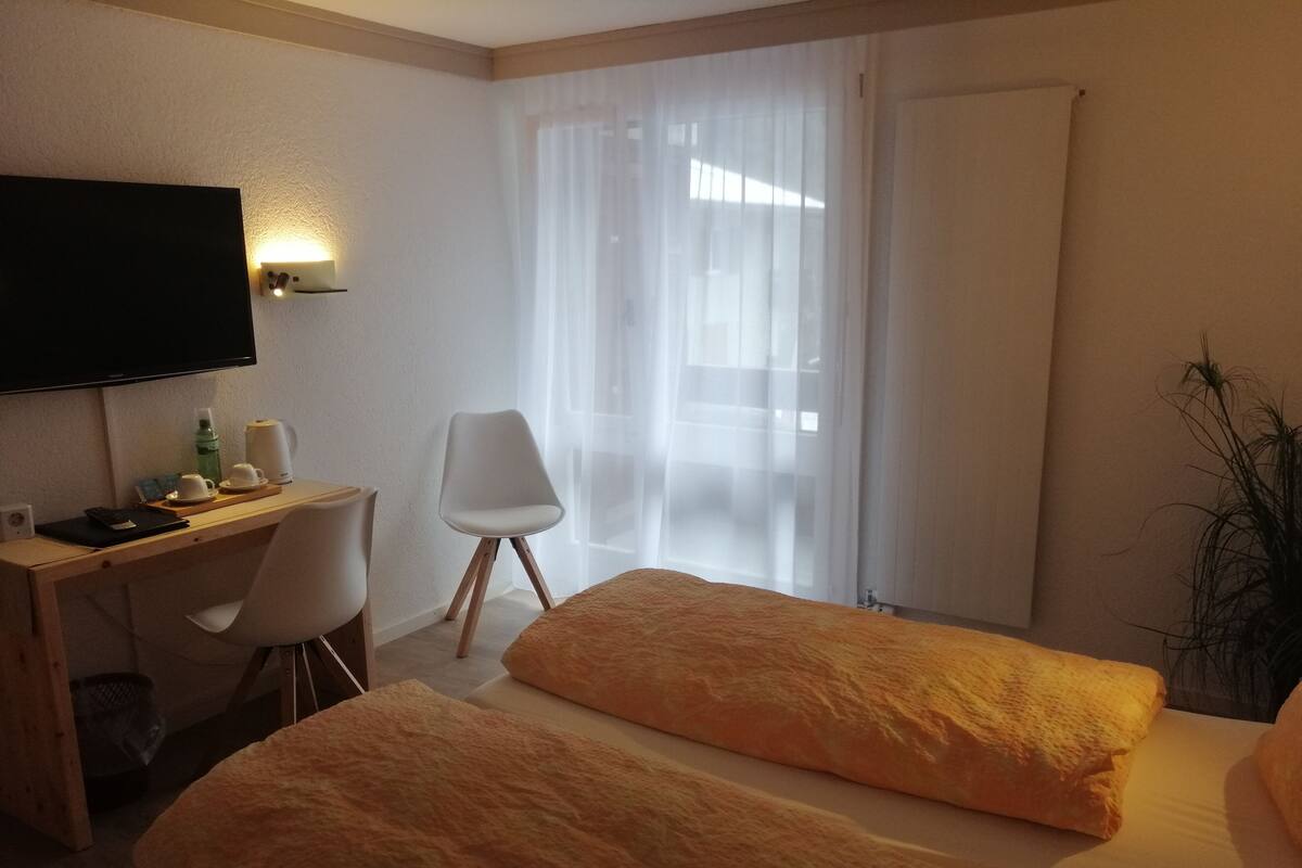 Quarto de hotel com cama, escrivaninha, cadeira, televisão e janela com cortinas leves.