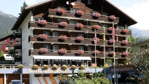 Un hotel de varios pisos con balcones decorados con macetas de flores y un letrero que indica 'HOTEL RESIDENCE', con montañas al fondo.