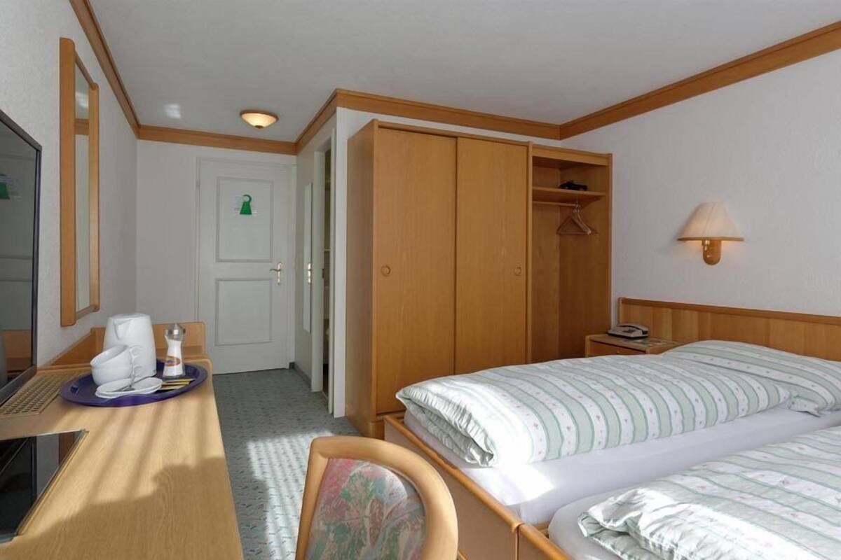 Quarto de hotel com uma cama, uma escrivaninha com cadeira, um guarda-roupa, uma mesinha de cabeceira e uma luminária.