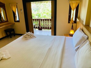 A hotel room with a large bed, a small table, a chair, and a balcony with a view of greenery.