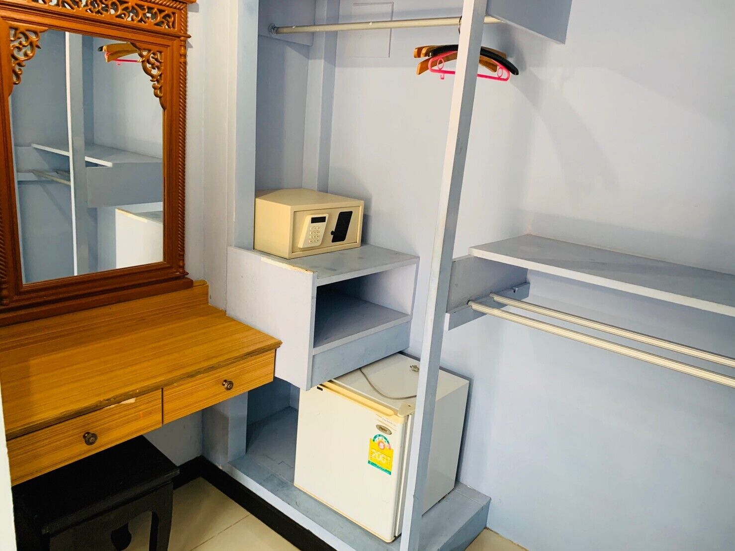 A room with a wooden desk, a mirror, a small refrigerator, and a wardrobe with shelves.