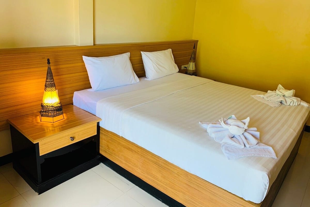 A neatly made bed with white linens and pillows, a wooden headboard, a bedside table with a lamp, and a folded towel on the bed.
