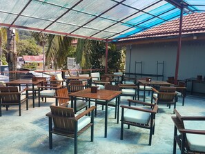 An outdoor cafe with wooden tables and chairs, a glass roof, and a view of a red-roofed building in the background.