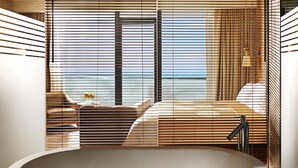 A modern bathroom with a large bathtub, wooden blinds, and a view of the ocean.