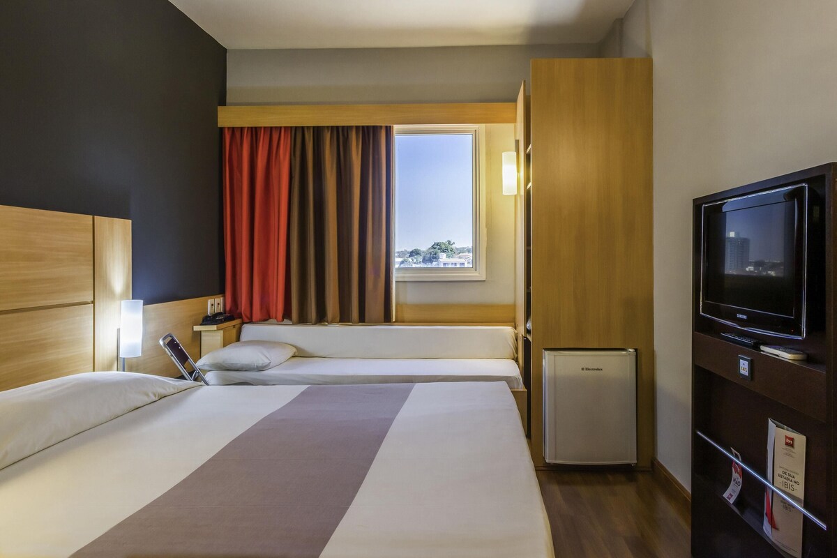 A hotel room with two beds, a television, a mini-fridge, and a window with curtains.
