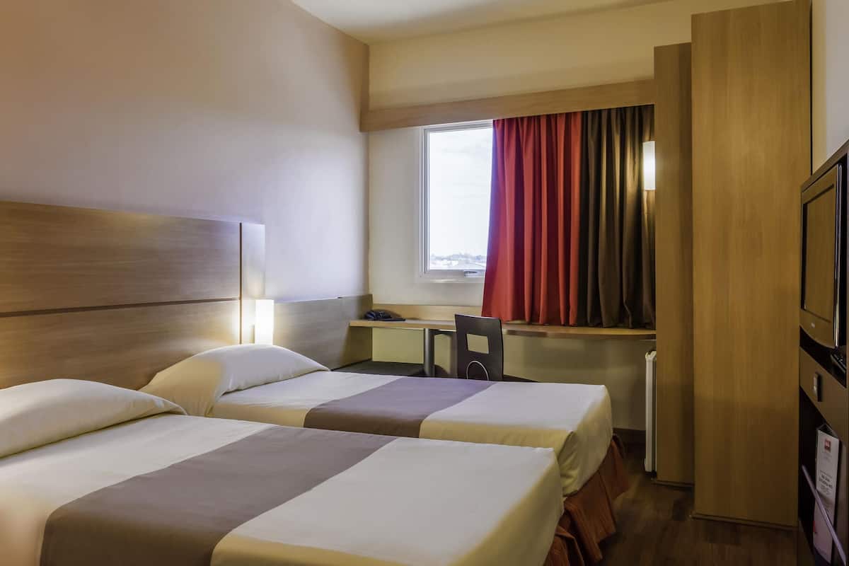 Une chambre d’hôtel avec deux lits, un bureau, une chaise, une télévision et une fenêtre avec des rideaux.