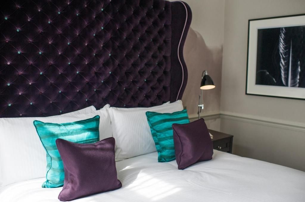 Una cama con cabecera capitonada, ropa de cama blanca y almohadas turquesa y moradas.