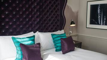 Una cama con cabecera capitonada, ropa de cama blanca y almohadas turquesa y moradas.
