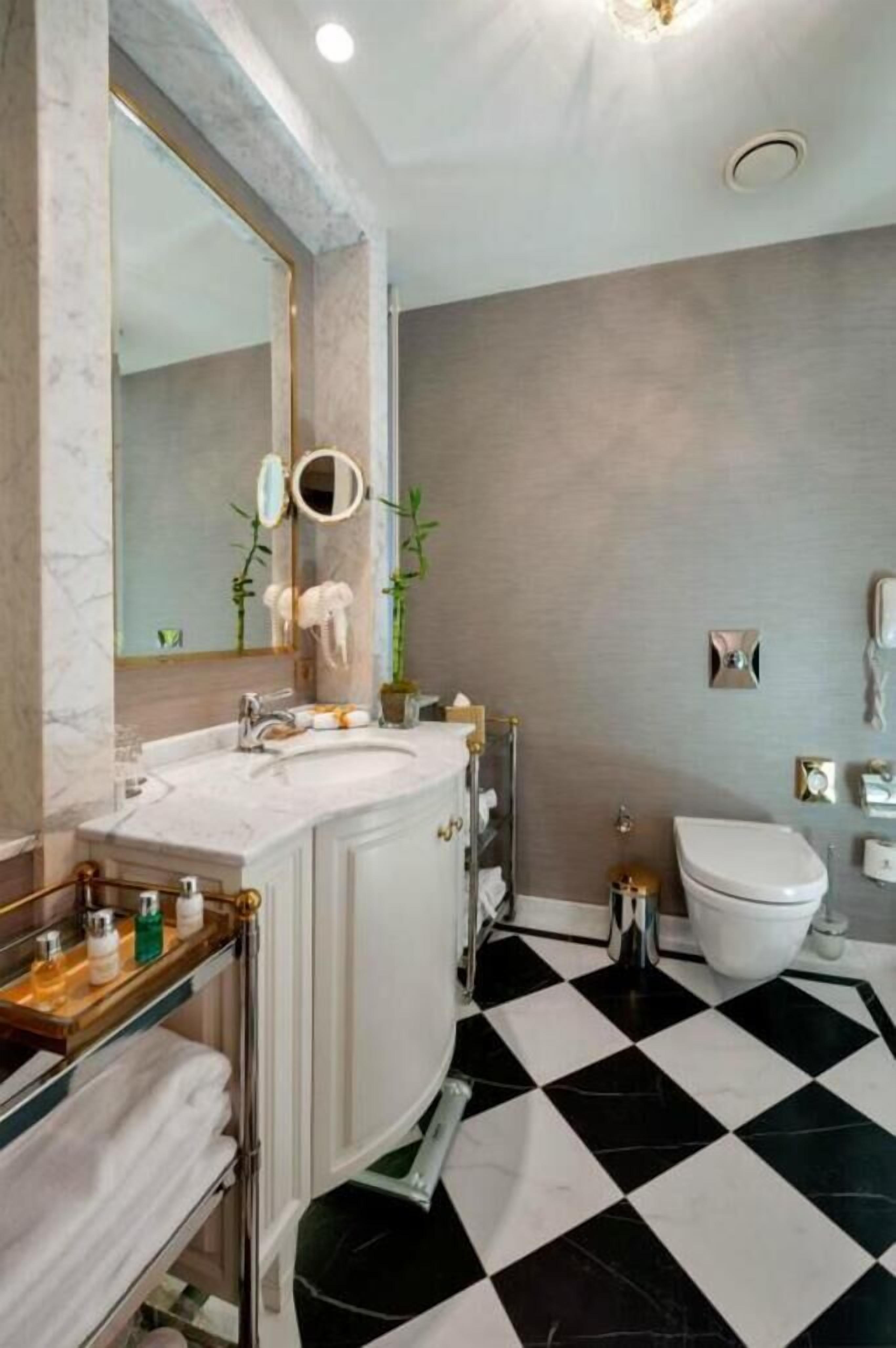 Een badkamer met een marmeren wastafelmeubel, een zwart-wit geruite vloer, een toilet en een spiegel met gouden lijst.