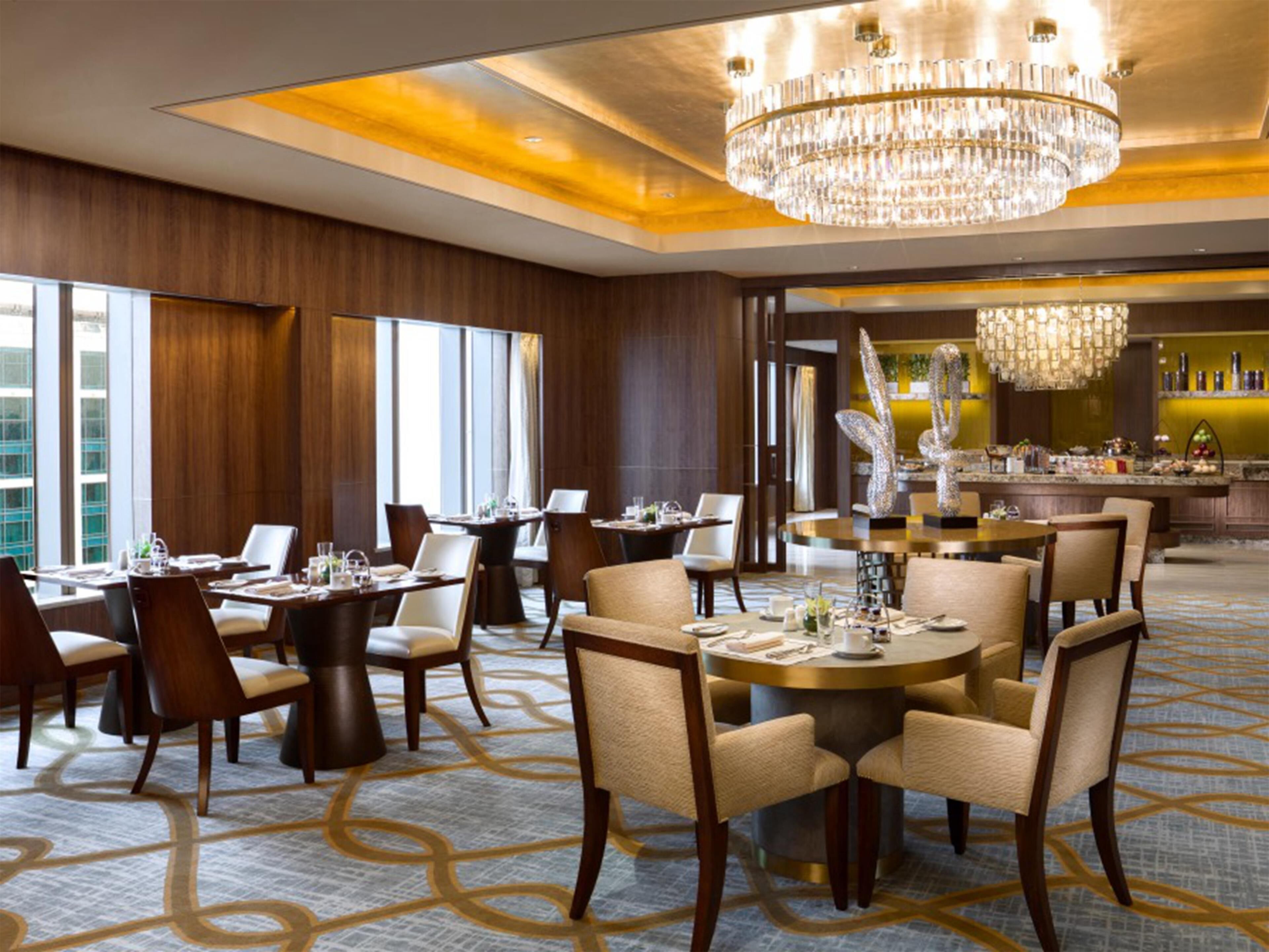 A modern hotel dining area with multiple tables and chairs, a large chandelier, and a bar area with a decorative sculpture.