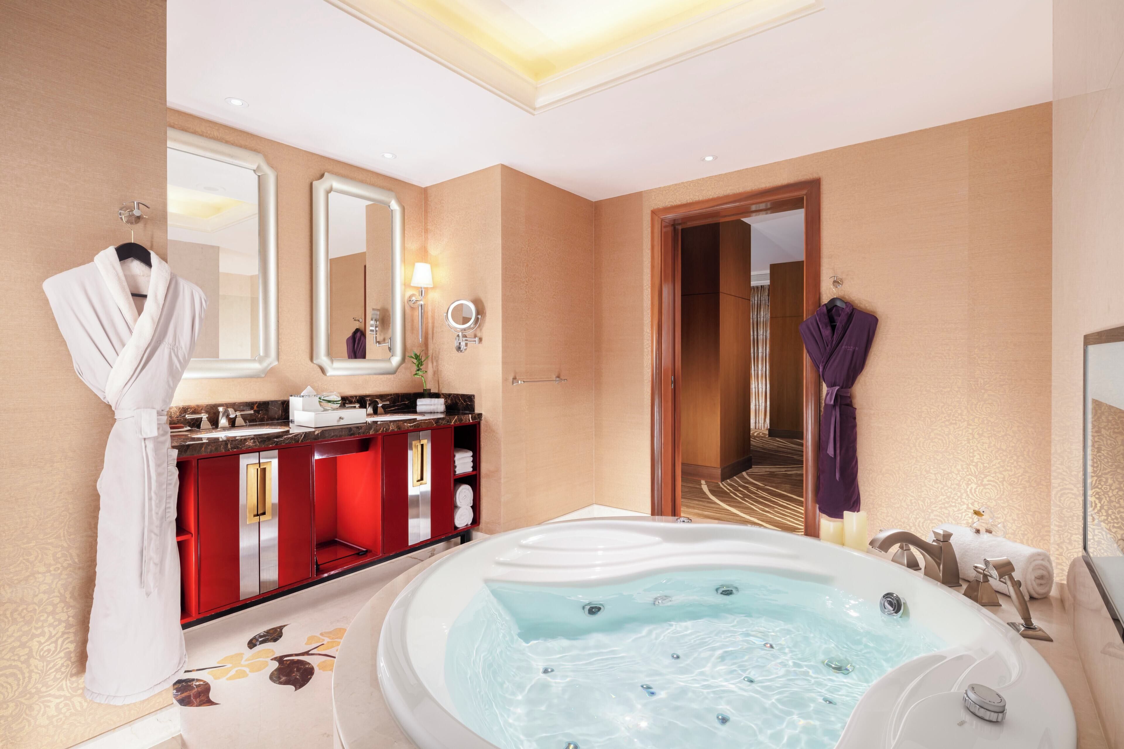 A luxurious bathroom with a large bathtub, a vanity with red cabinets, and two robes hanging on hooks.