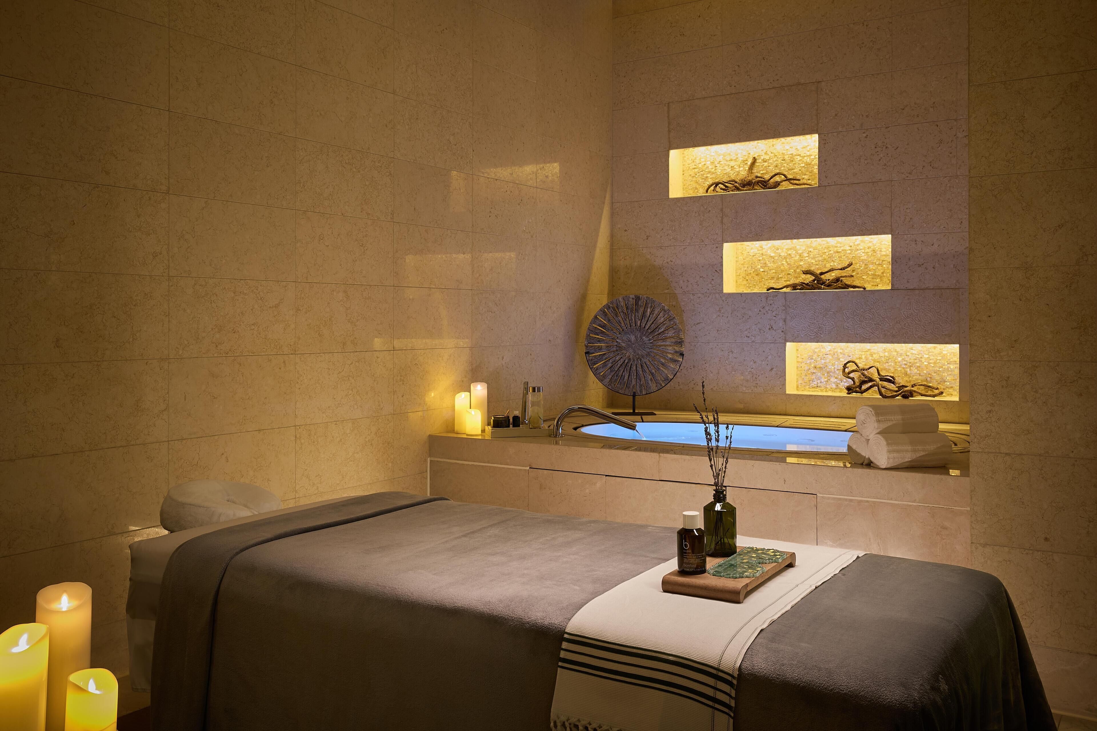 Een wellnessruimte met een massagetafel, kaarsen, een wastafel en wandplanken met decoratieve objecten.