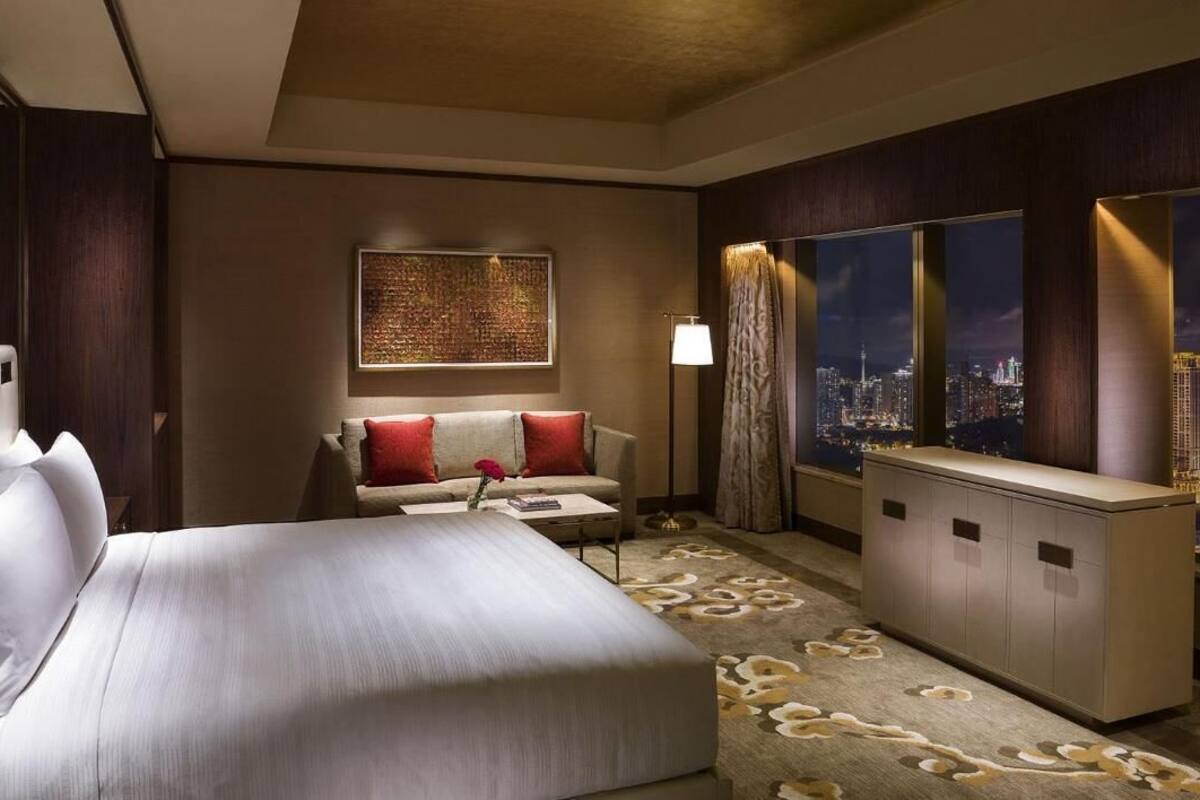 Habitación de hotel con una cama grande, un sofá con cojines rojos, un escritorio, un ventanal con vistas a la ciudad y un elemento decorativo en la pared.