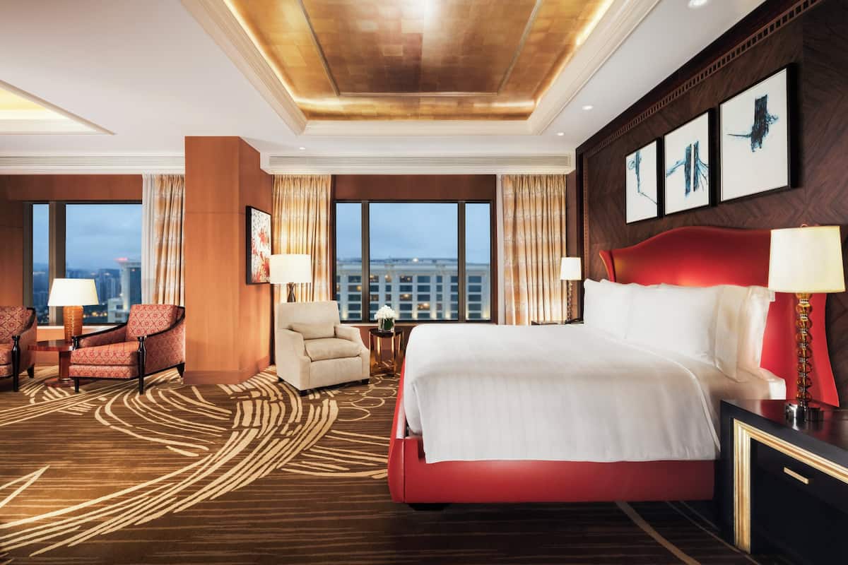 Habitación de hotel con una cama grande, dos sillones, una mesita y vistas a la ciudad a través de amplios ventanales.