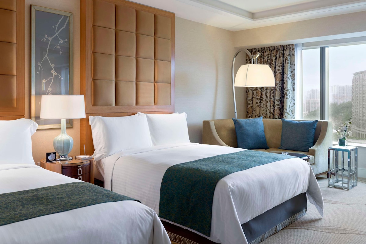 A hotel room with two beds, a large window with a view of buildings, a modern lamp, and a decorative wall panel.