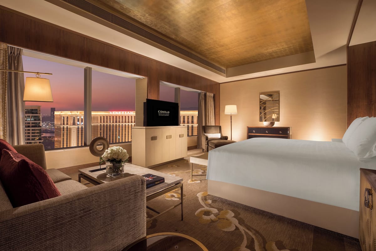 Habitación de hotel con cama, televisor, escritorio con lámpara, una silla y vista a un paisaje urbano al atardecer.