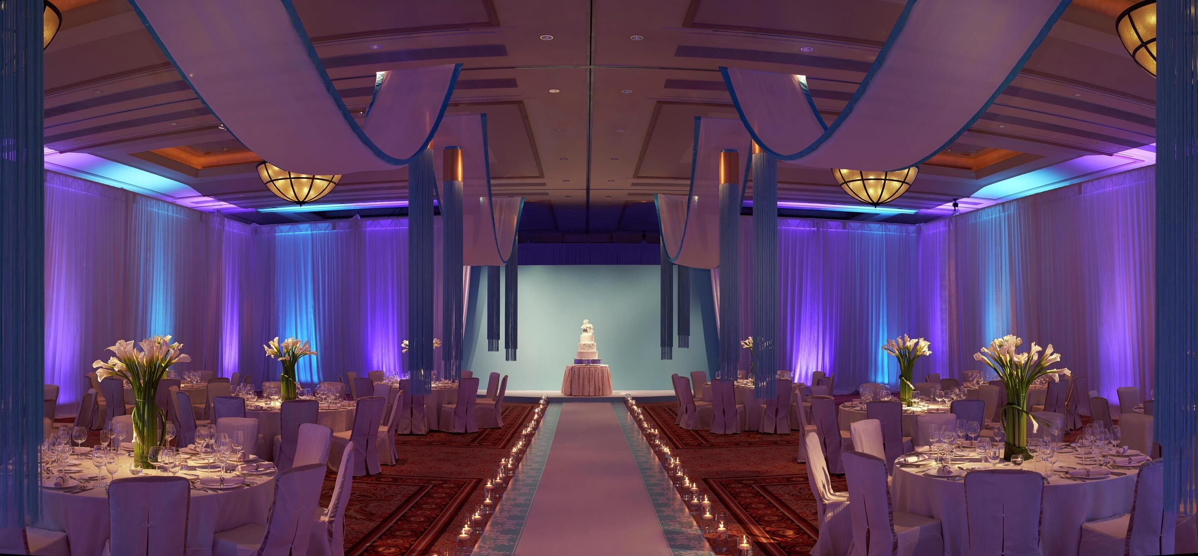 Un salón de banquetes con mesas redondas, sillas blancas y un escenario con un pastel.