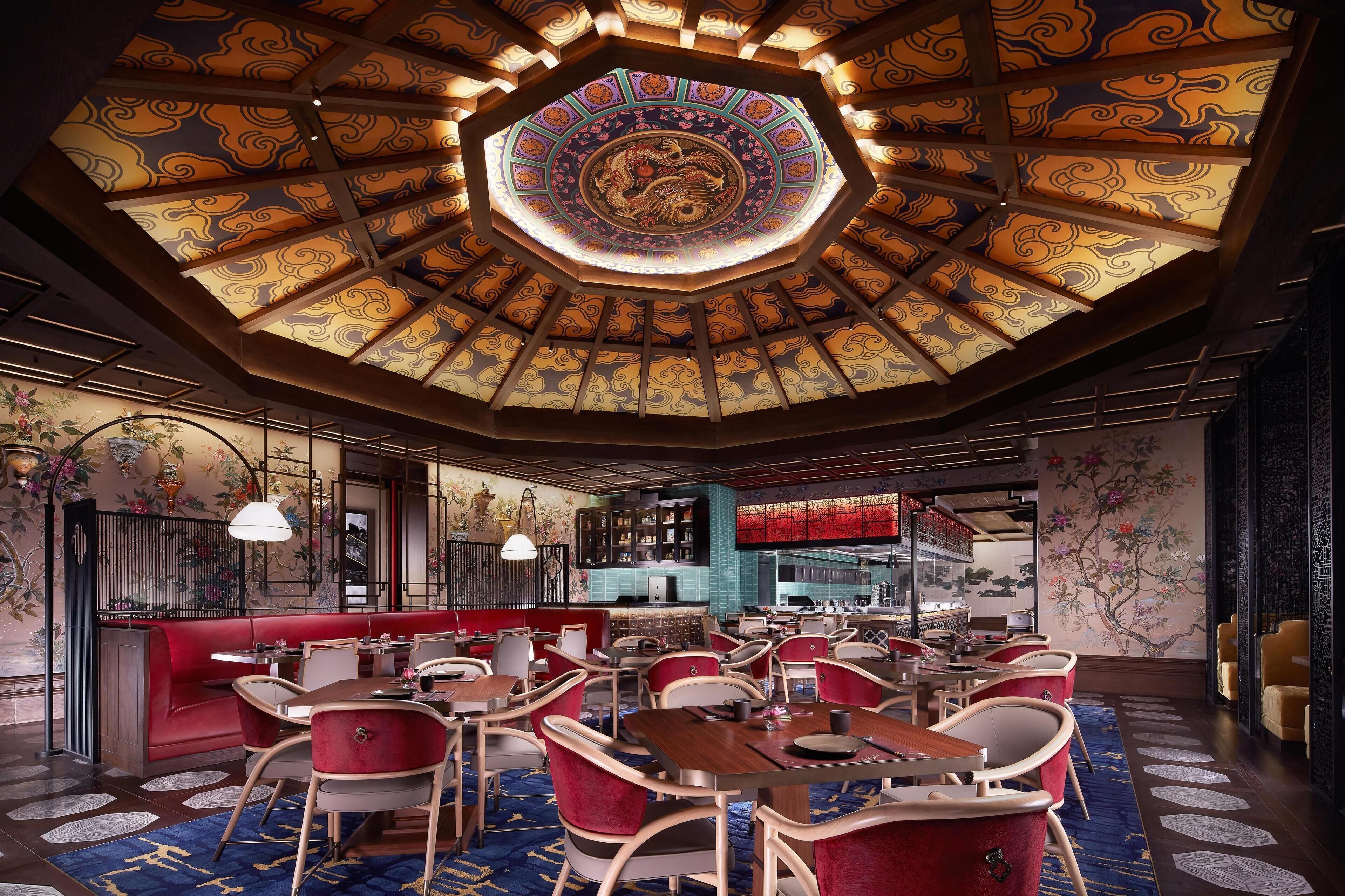 A restaurant with a traditional design, featuring a central octagonal ceiling with intricate patterns and a large circular emblem. Tables are arranged around a red seating area, and the walls are adorned with detailed murals.