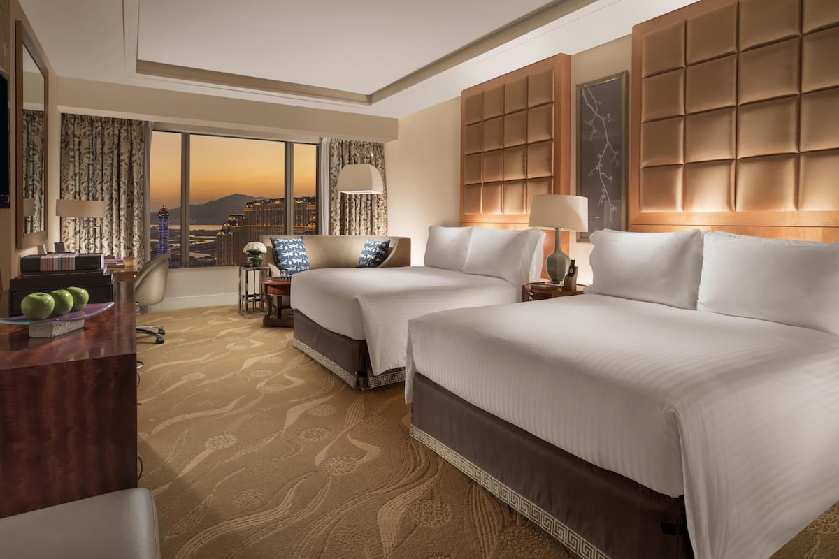 Habitación de hotel con dos camas, un escritorio, una silla, un ventanal grande con vistas a la ciudad y un cabecero decorativo.
