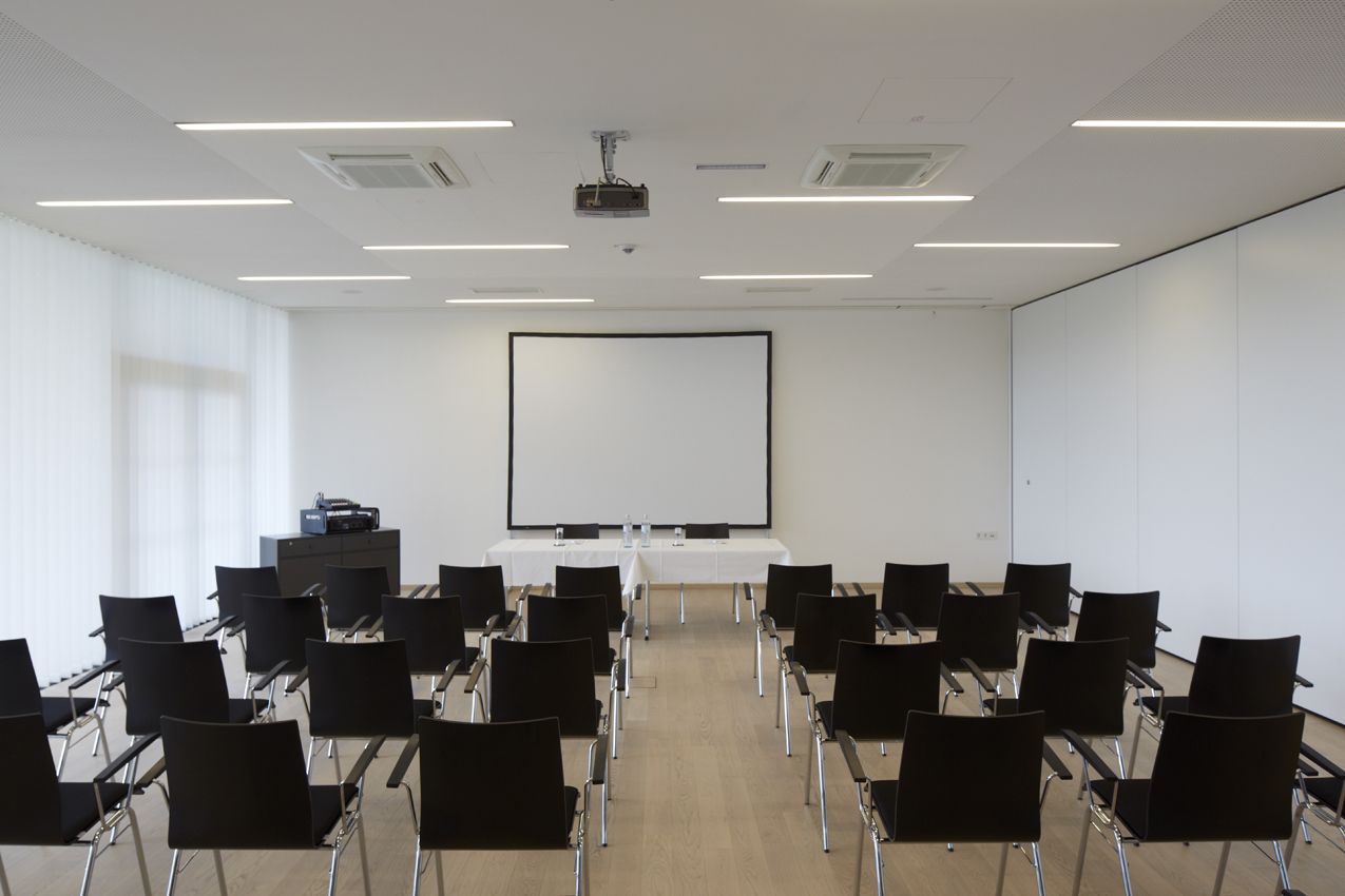 Ein Konferenzraum mit einer großen weißen Leinwand, Reihen schwarzer Stühle und einem an der Decke befestigten Projektor.