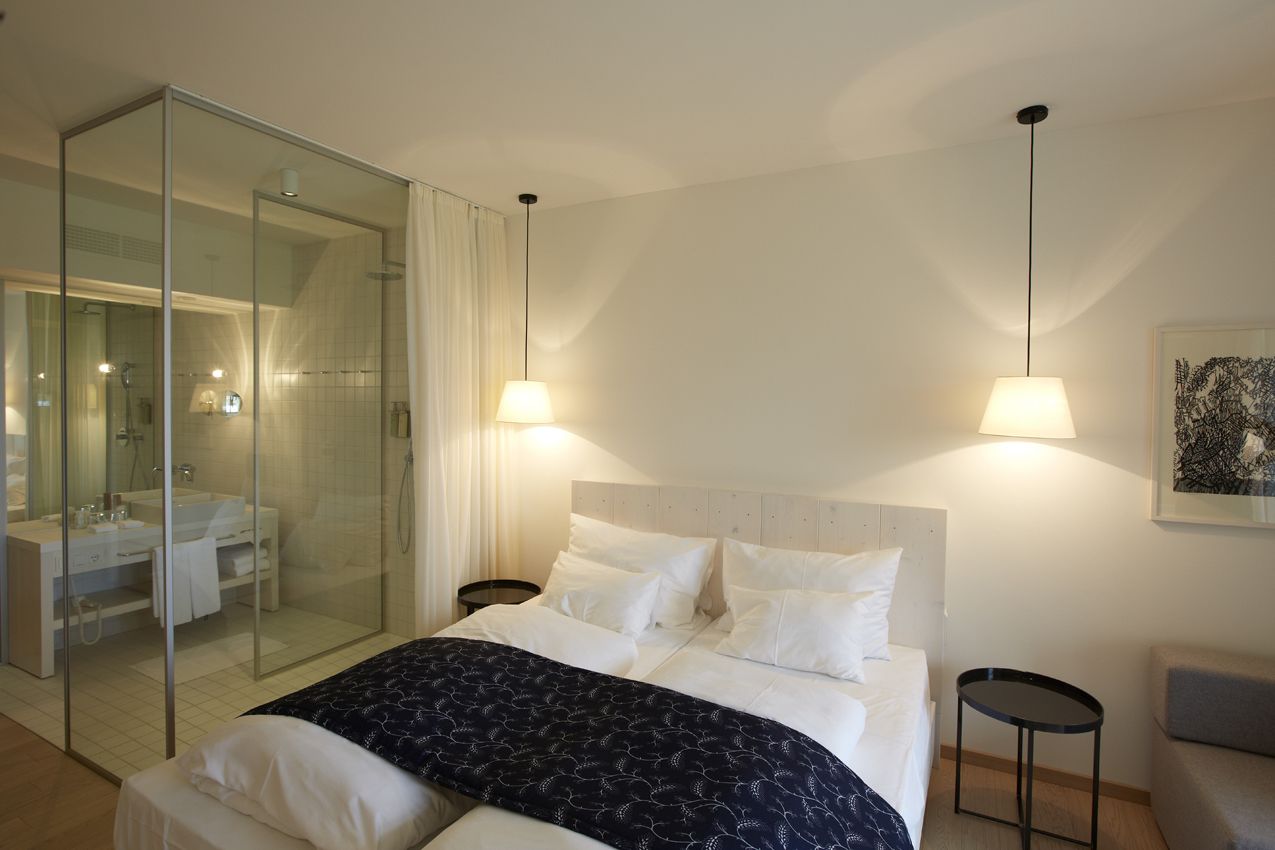 Habitación de hotel moderna con ducha de vidrio y cama con ropa blanca, además de dos lámparas de noche.