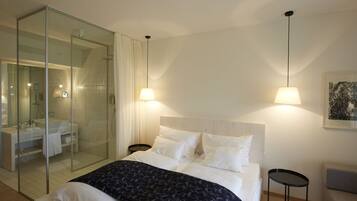 Habitación de hotel moderna con ducha de vidrio y cama con ropa blanca, además de dos lámparas de noche.