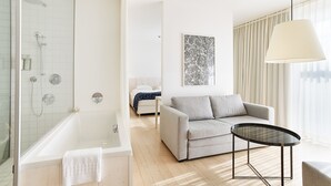 A modern bathroom with a bathtub, shower, and toilet. There is a grey sofa and a small round table in the living area. - Loisium Wine & Spa Hotel Südsteiermark  (Ehrenhausen an der Weinstraße)