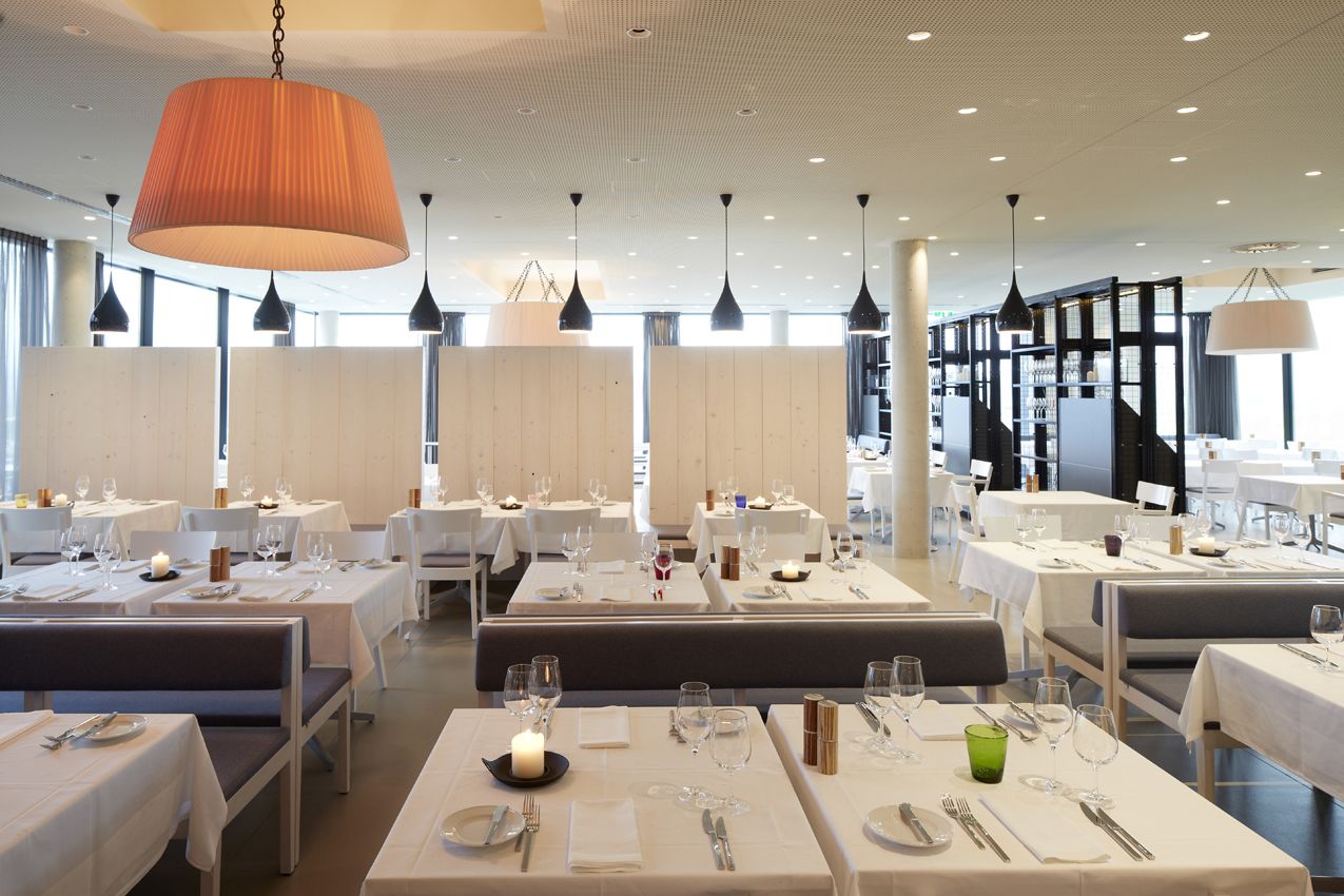 Een modern restaurant met witte tafelkleden, zwarte hanglampen en een grote, oranje hangende lamp.