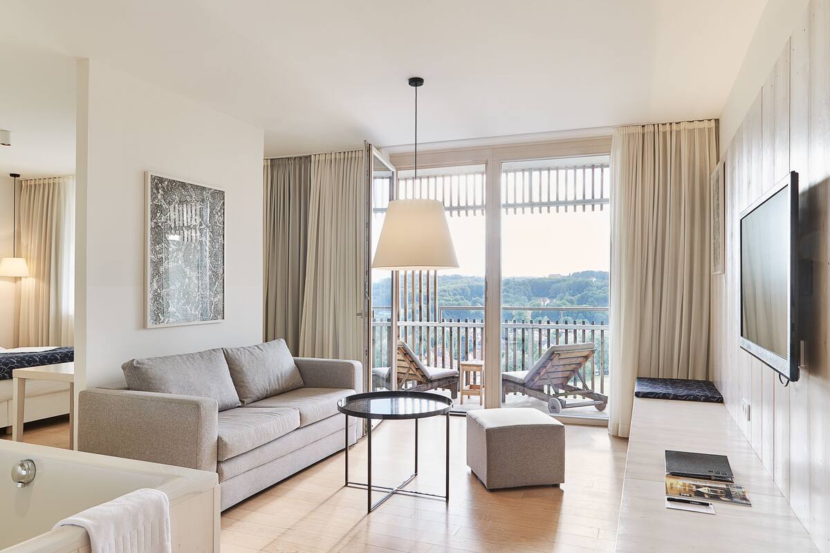 Ein modernes Hotelzimmer mit einer Couch, einem runden Couchtisch, einem Fernseher und einem Balkon mit Blick.