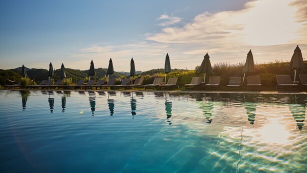 A clear swimming pool with lounge chairs and umbrellas, surrounded by greenery and hills. - Loisium Wine & Spa Hotel Südsteiermark  (Ehrenhausen an der Weinstraße)
