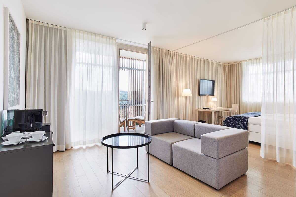 Ein modernes Hotelzimmer mit einem grauen Ecksofa, einem kleinen runden Tisch, einem Fernseher und einem Balkon mit Vorhängen.