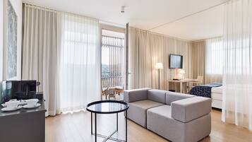 Ein modernes Hotelzimmer mit einem grauen Ecksofa, einem kleinen runden Tisch, einem Fernseher und einem Balkon mit Vorhängen.