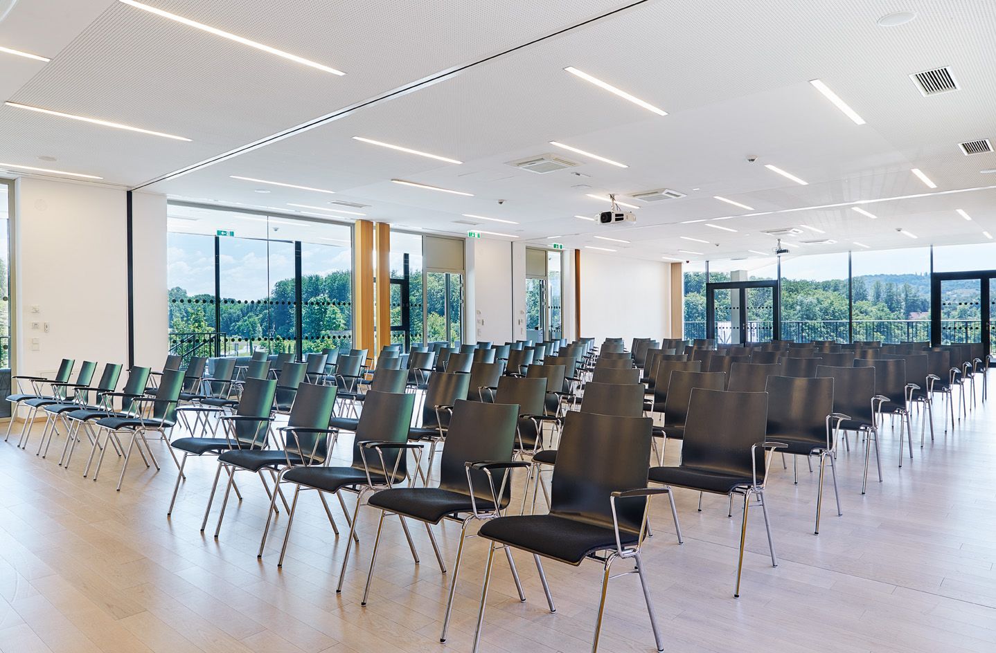 Un salón de conferencias con hileras de sillas, ventanas amplias y un techo con luminarias lineales.