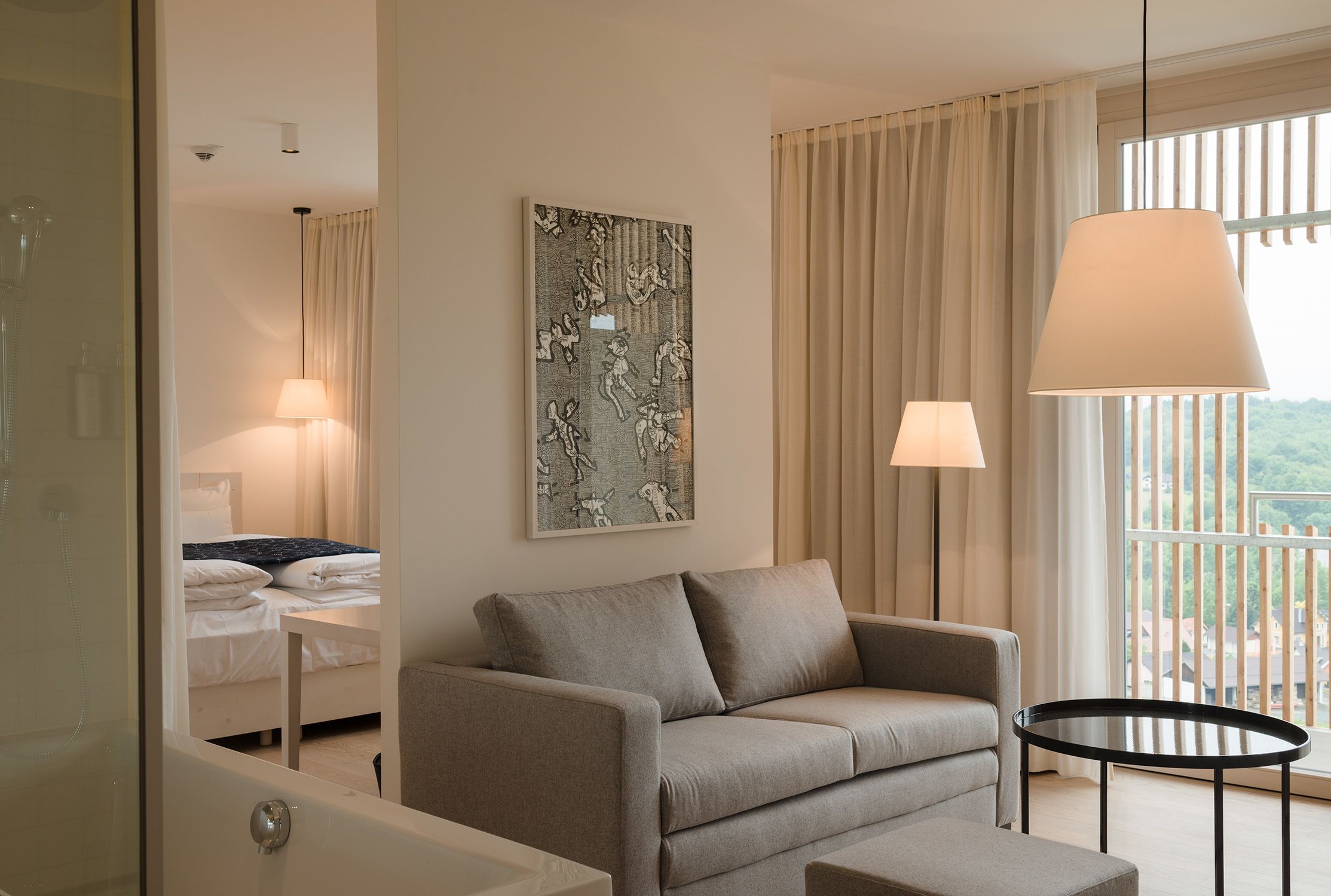 A modern hotel room with a sofa, a small round table, a painting on the wall, and a large window with curtains.